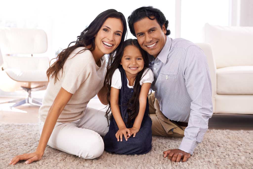 mother father and young child sitting on carpeted living room floor smiling