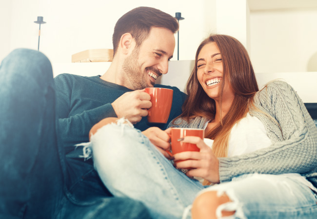 Man and woman with dental implants smile as they cuddle on a couch holding orange mugs